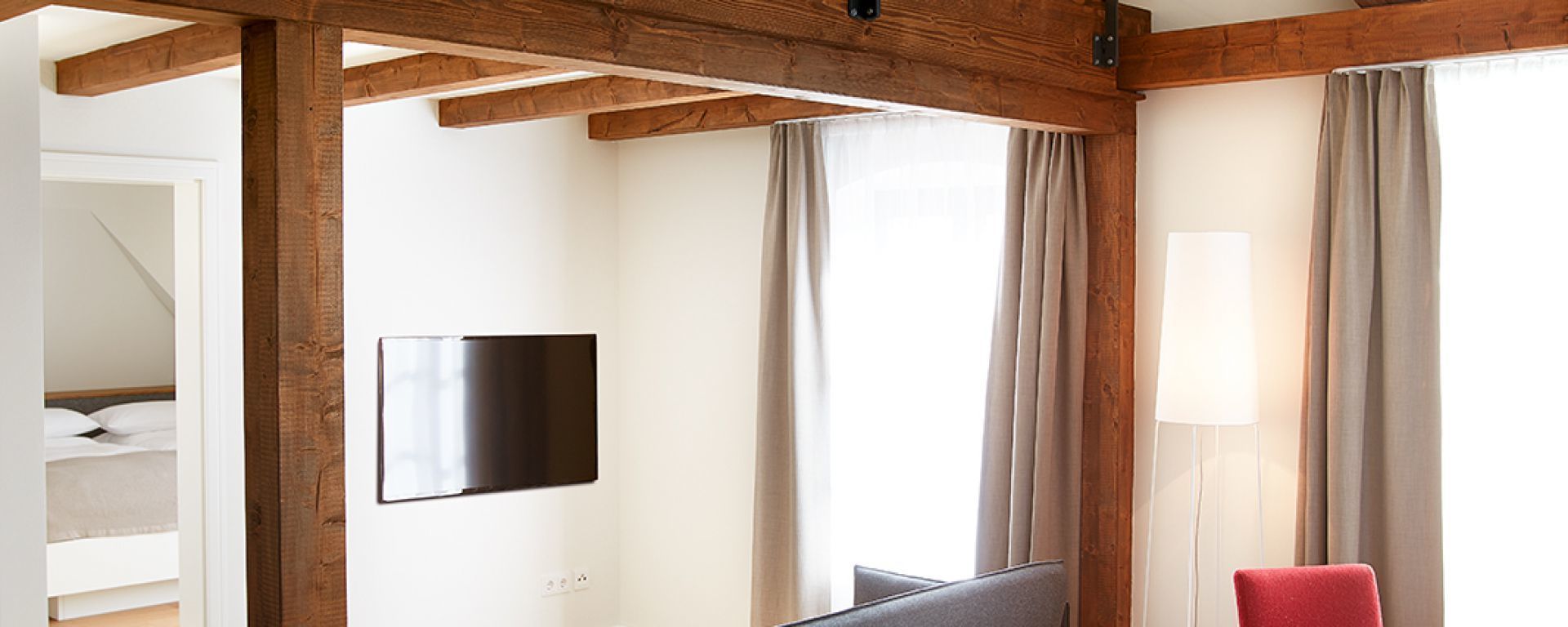A hotel room with a wooden ceiling, a flat-screen TV mounted on the wall, a bed with white bedding, and a red chair.