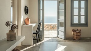 A room with a stone wall, a white bench, a chair, a wicker basket, and a view of the sea through an open door.