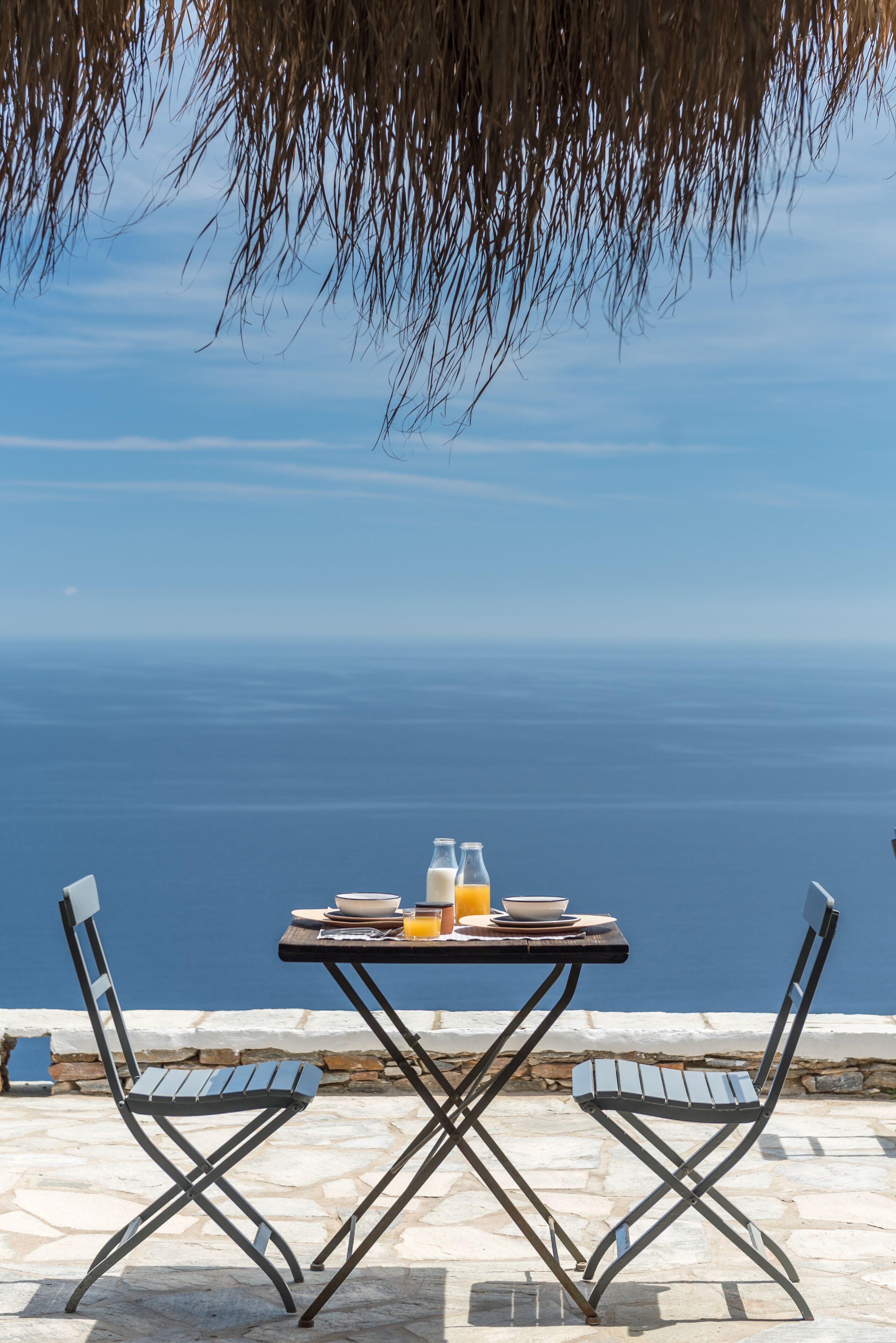 A table with two chairs, a glass of orange juice, and a bowl on a stone-paved area with a view of the sea.