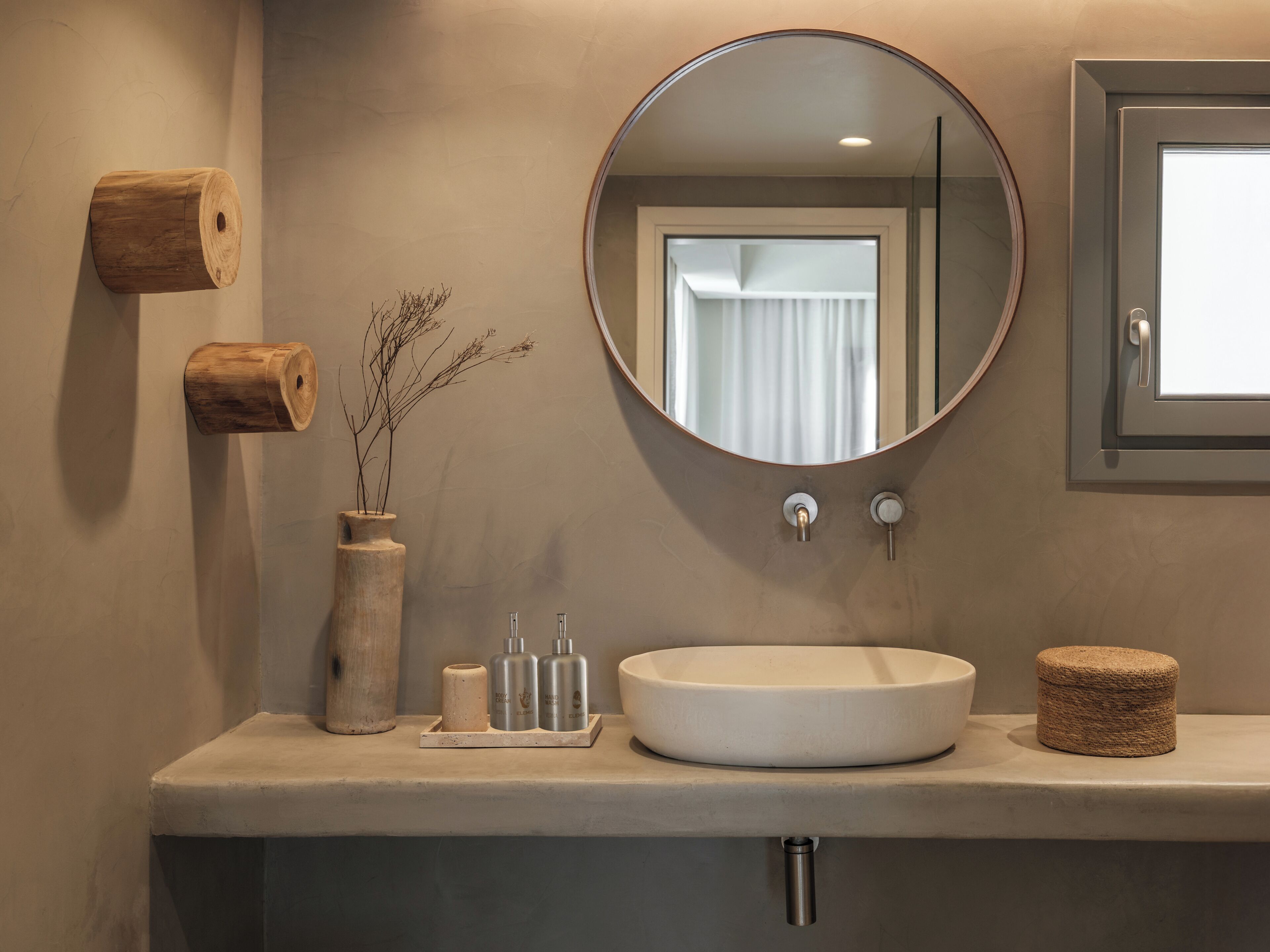 Een badkamer met een ronde spiegel, een wastafel, een vaas met gedroogde planten en een klein houten krukje.