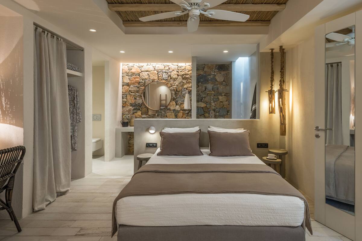 Un dormitorio con una cama grande, un ventilador de techo, una pared de piedra, un espejo y una mesita de noche con lámpara.