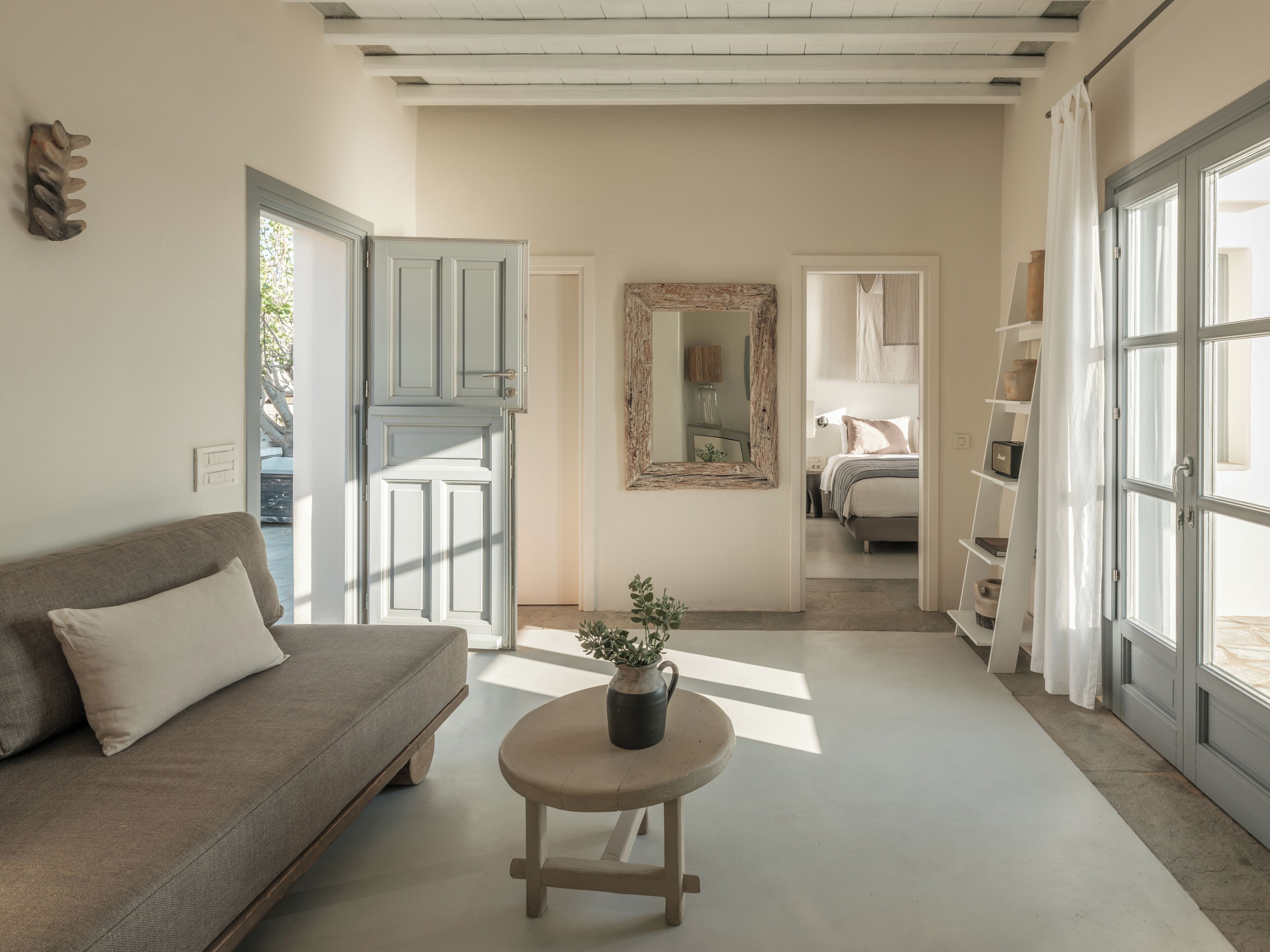 Una habitación con un sofá, una mesita redonda con una planta, un espejo y una cama al fondo.