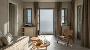 A spacious room with a large window offering a view of the sea, a comfortable sofa, a wooden chair, and a small table with a vase.