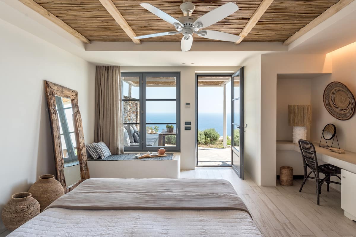 Un dormitorio con una cama grande, un ventilador de techo, vista al mar a través de una puerta abierta y un escritorio con una silla.