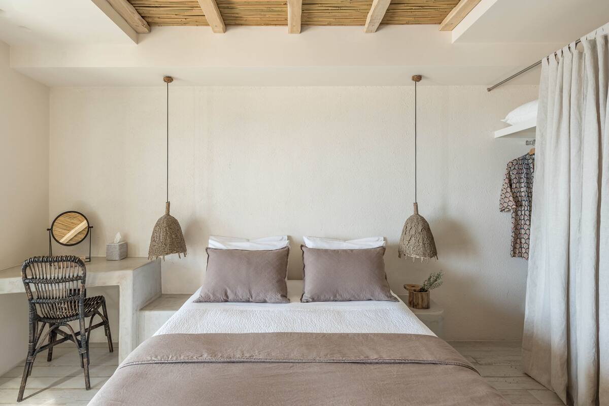 Un dormitorio con cama, dos lámparas colgantes, una silla, una mesita de noche y una ventana con cortinas.