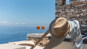 A beachside lounge chair with a straw hat, two glasses of orange drink, and a stone wall backdrop.