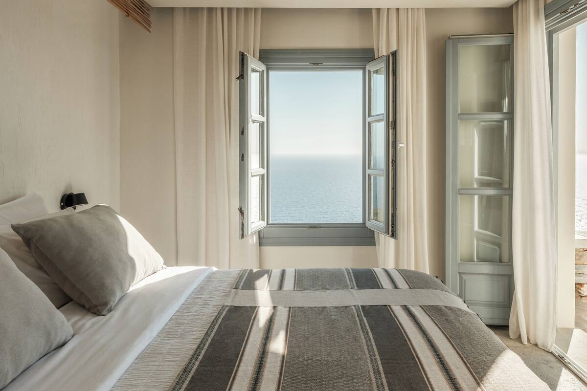 Un dormitorio con cama, almohadas, una mesita de noche, una ventana con cortinas y vistas al mar.