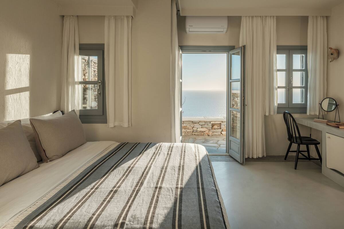 Un dormitorio con cama, un escritorio con silla y vista al mar a través de una puerta abierta.