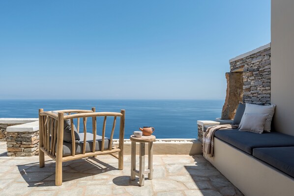 Una silla de madera y una mesita con una tetera en un patio de piedra con vista al mar.