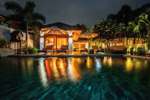 A poolside area with lounge chairs and umbrellas, surrounded by palm trees and a building with warm lighting.