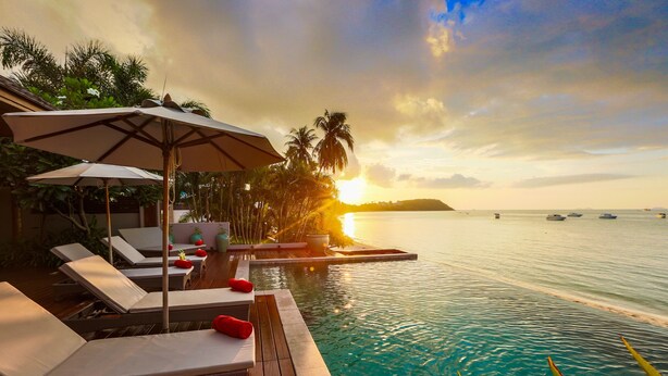 Zona de piscina con sillones y sombrillas, con vista al mar tranquilo y al atardecer.