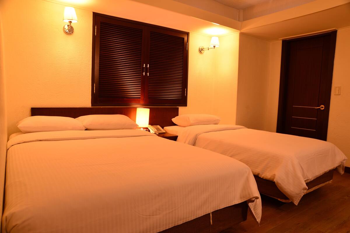 A hotel room with two beds, a wooden headboard, a bedside table with a lamp, and a door.