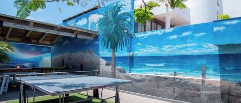 An outdoor area with a ping pong table, a mural of a beach scene, and a palm tree.