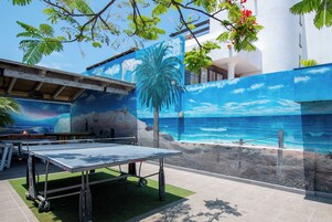 Área externa com mesa de pingue-pongue, um mural de uma praia e uma palmeira.