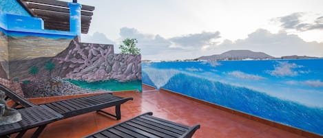 Una terraza con sillones de madera, un muro de piedra con un mural y vista al océano.