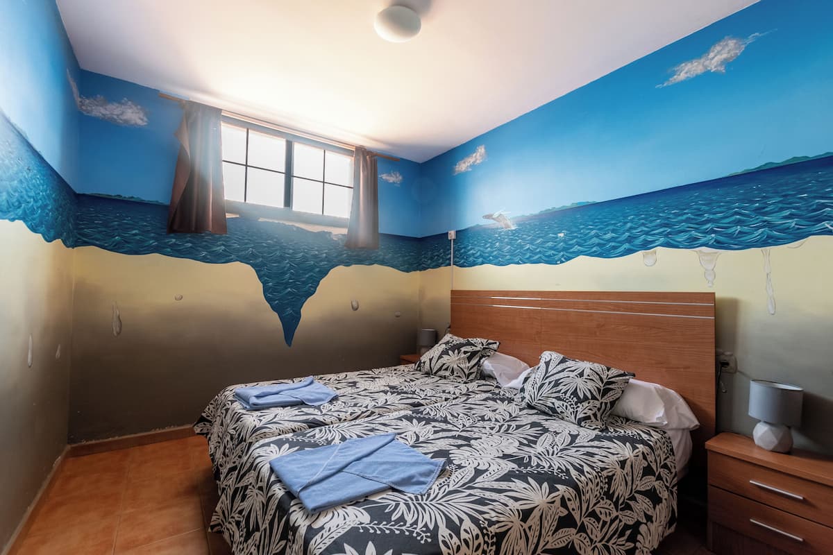 A bedroom with a mural of the ocean on the walls, a bed with a patterned bedspread, a window with curtains, and a bedside table with a lamp.