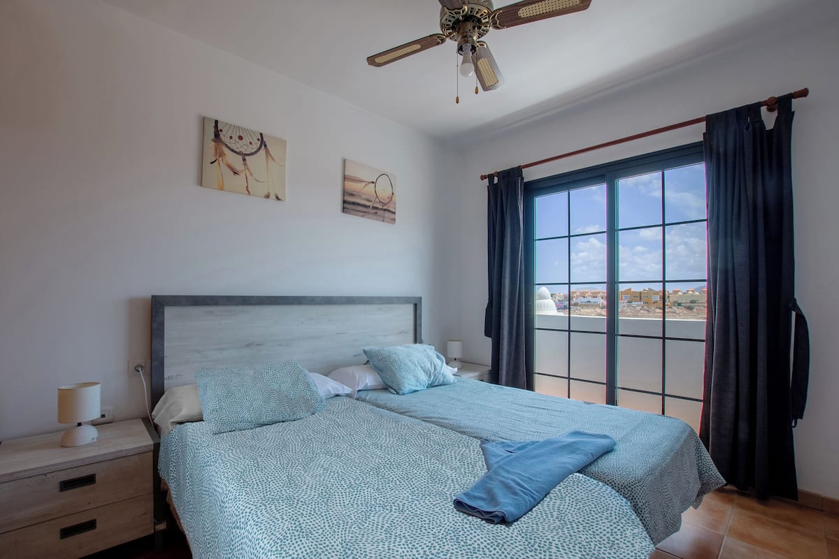 A bedroom with a large bed, a nightstand, a ceiling fan, and a window with curtains.