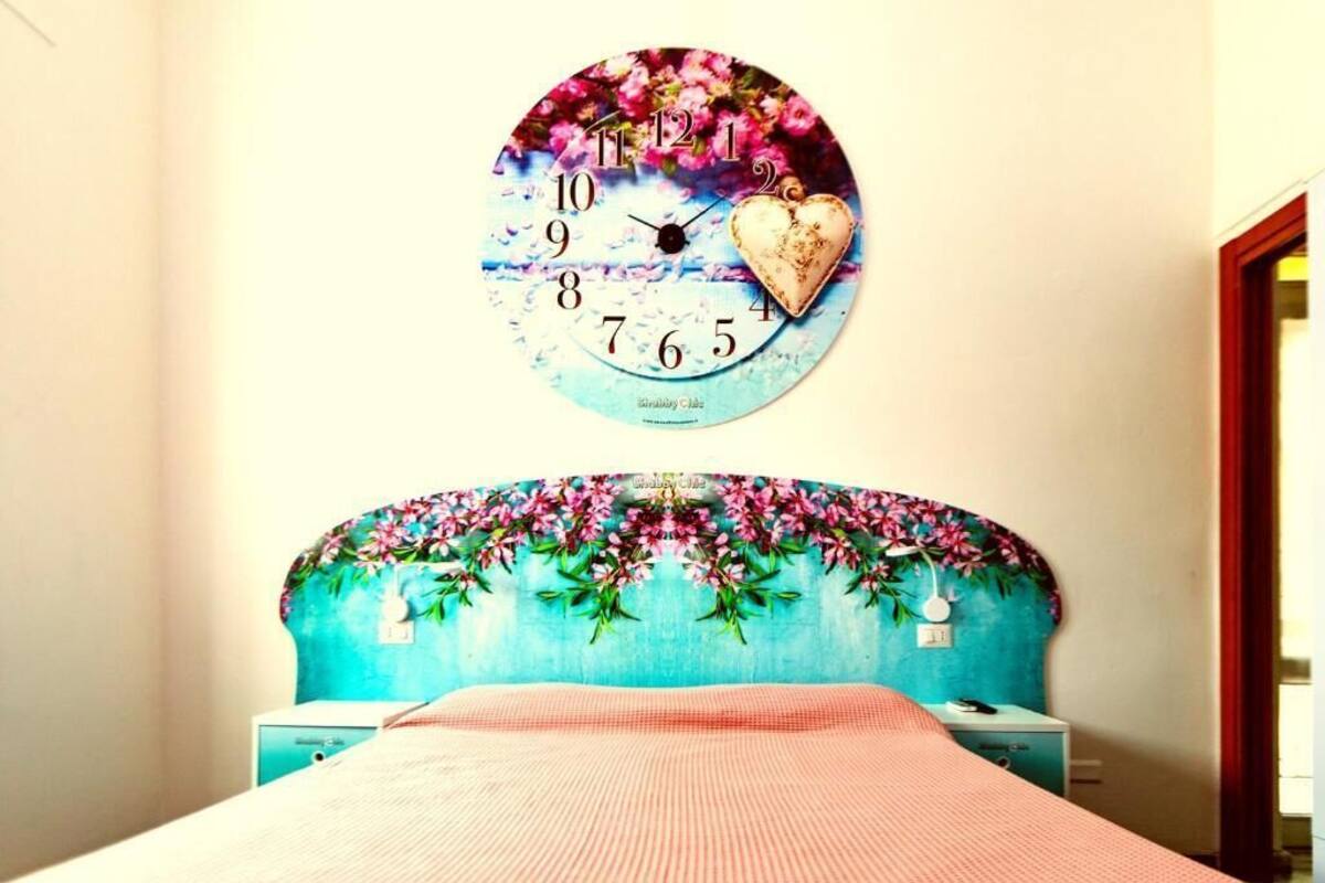 A bedroom with a floral headboard, a round wall clock, and a bed with a pink blanket.