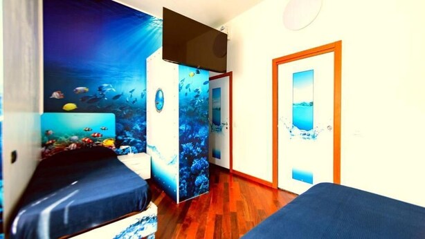 A room with a wooden floor, a bed with a blue bedspread, a wall with an underwater mural, and a door with a window. - Pavia AffittaCamere (Pavia)