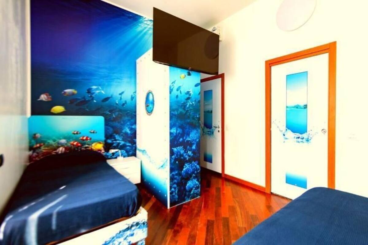 A room with a wooden floor, a bed with a blue bedspread, a wall with an underwater mural, and a door with a window.