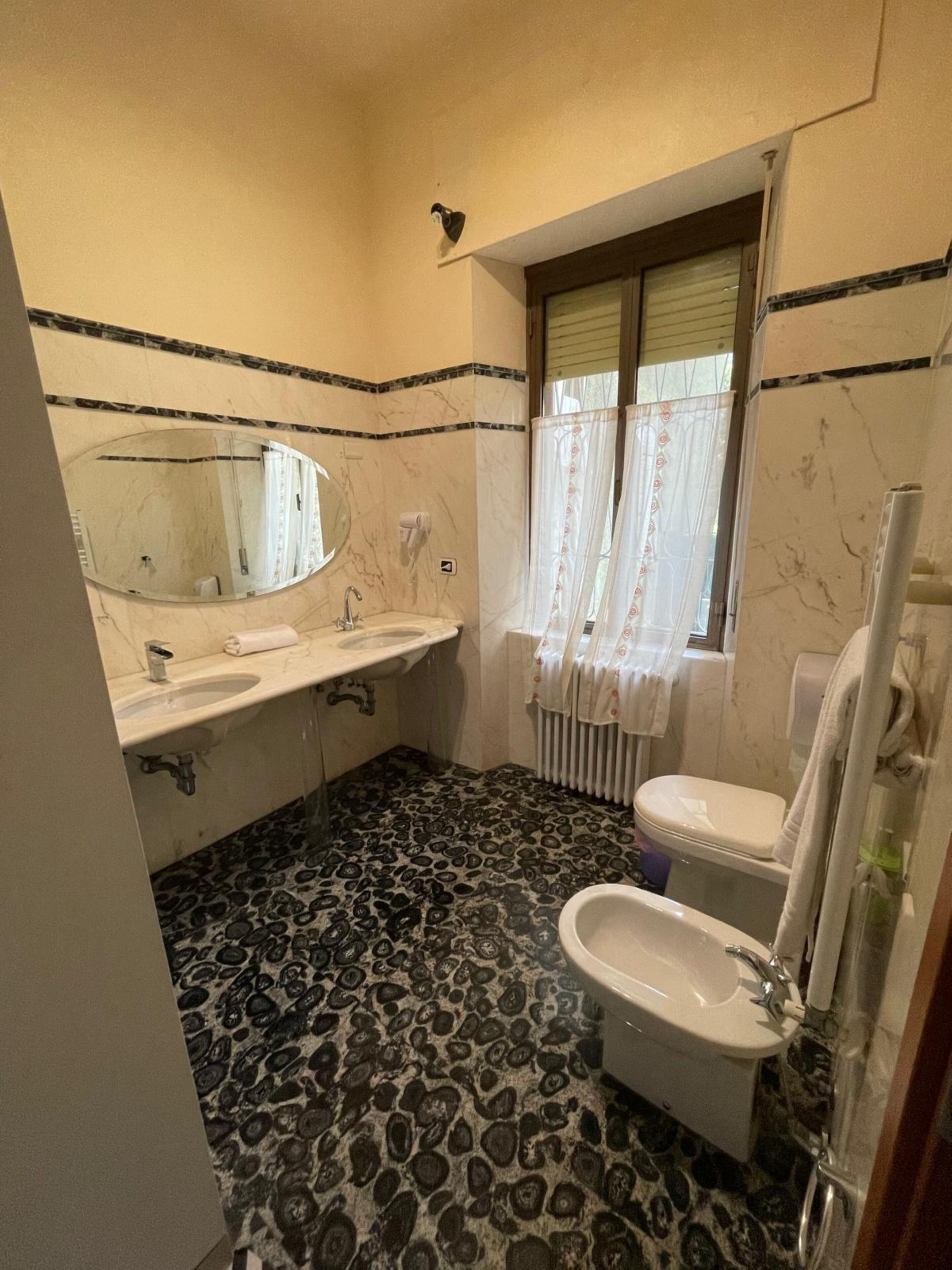 Ein Badezimmer mit zwei Waschbecken, einer Toilette, einem Bidet, einem Spiegel und einem fenster mit Vorhängen.