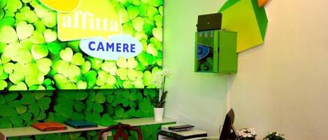 Una habitación con pared estampada de hojas verdes, una mesa de vidrio y un escritorio con una computadora y material de oficina.