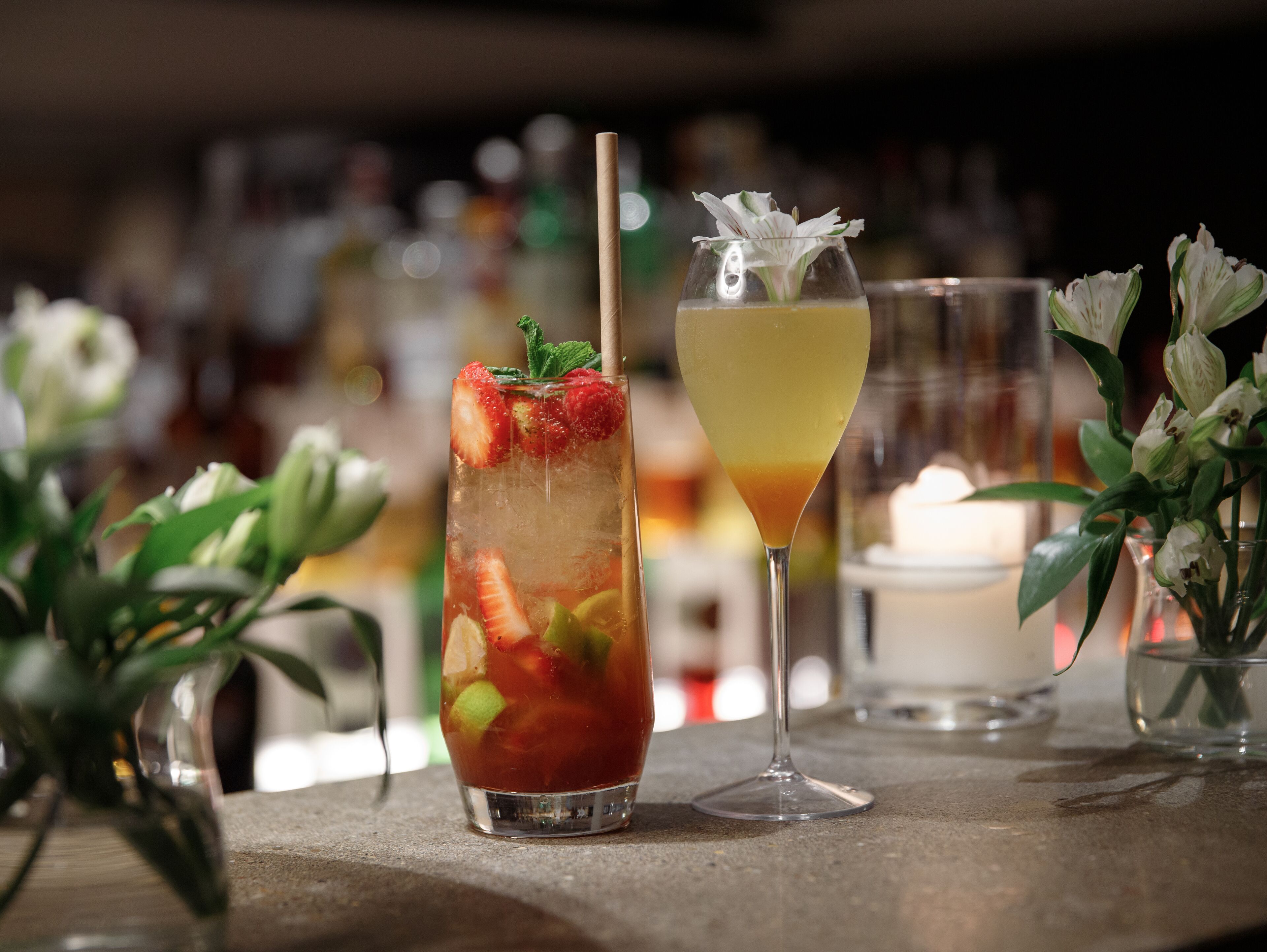 A bar setting with two drinks, one with strawberries and mint, and another with a yellow liquid and a flower garnish.