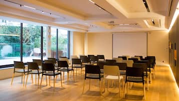 Un salón de conferencias con filas de sillas, una pantalla de proyector y grandes ventanales que dan a vistas de vegetación.