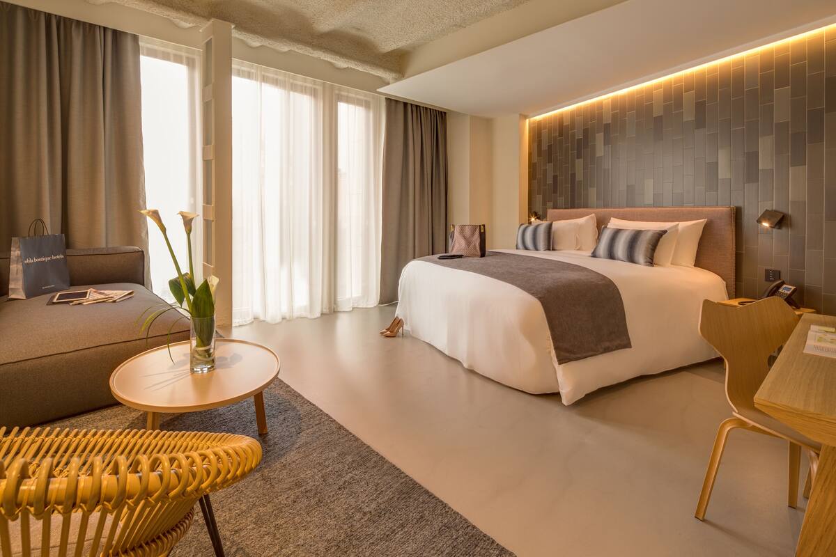 A modern hotel room with a large bed, a sofa, a small round table, and a chair.
