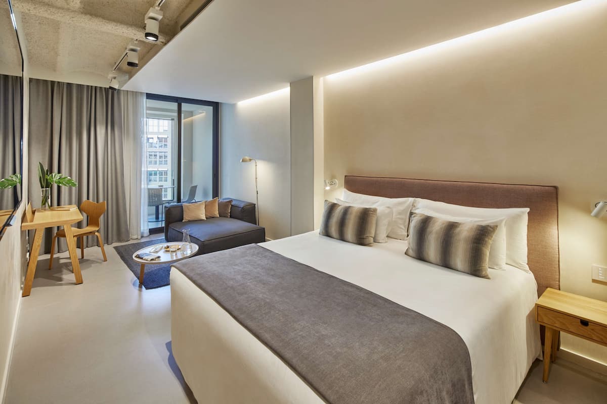 A modern hotel room with a large bed, a desk, a sofa, and a view of the city.