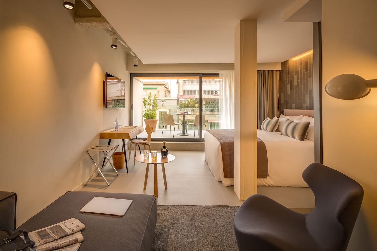 A modern hotel room with a bed, a desk, a chair, and a balcony with a view of the city.