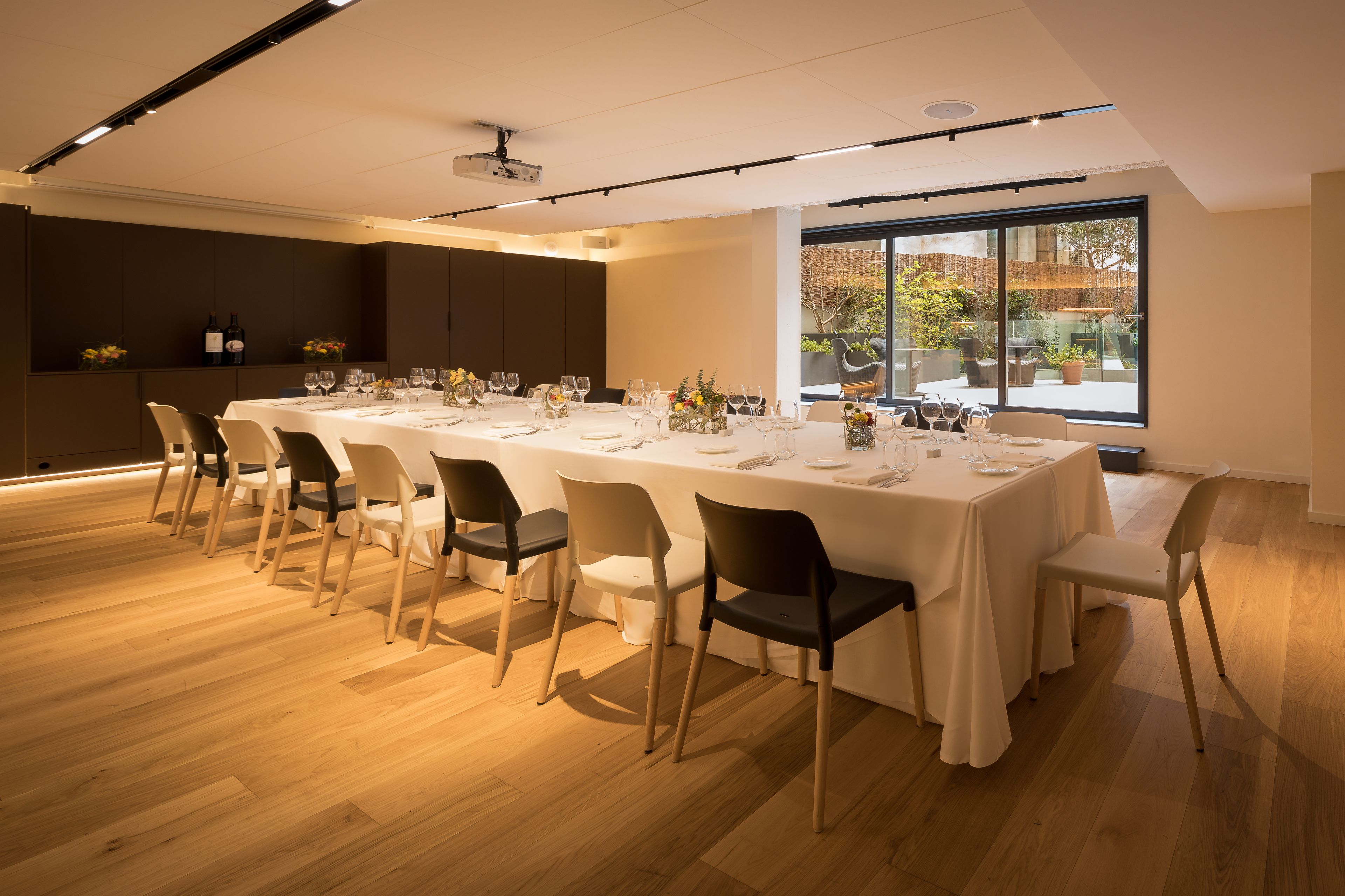 A modern dining area with a long table set for a meal, wooden flooring, and large windows offering a view of the outdoors.