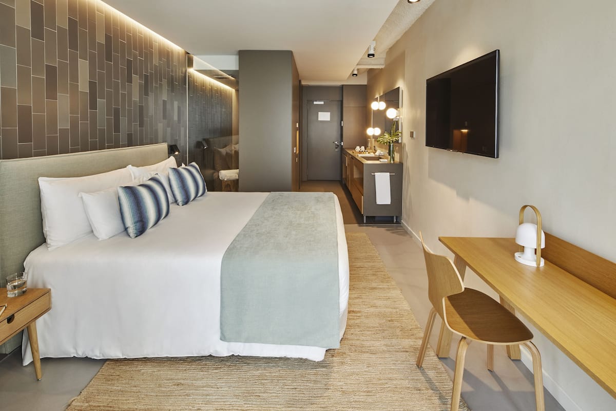 A modern hotel room with a large bed, a desk, a chair, a TV, and a bathroom visible in the background.
