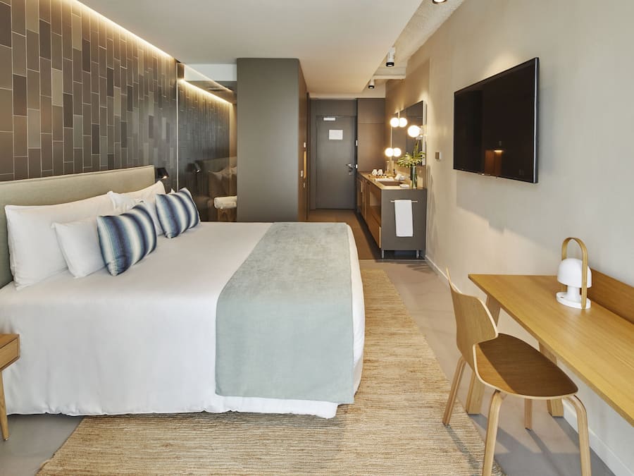 Quarto de hotel moderno com uma cama grande, uma escrivaninha, uma cadeira, uma televisão e um banheiro visível ao fundo.