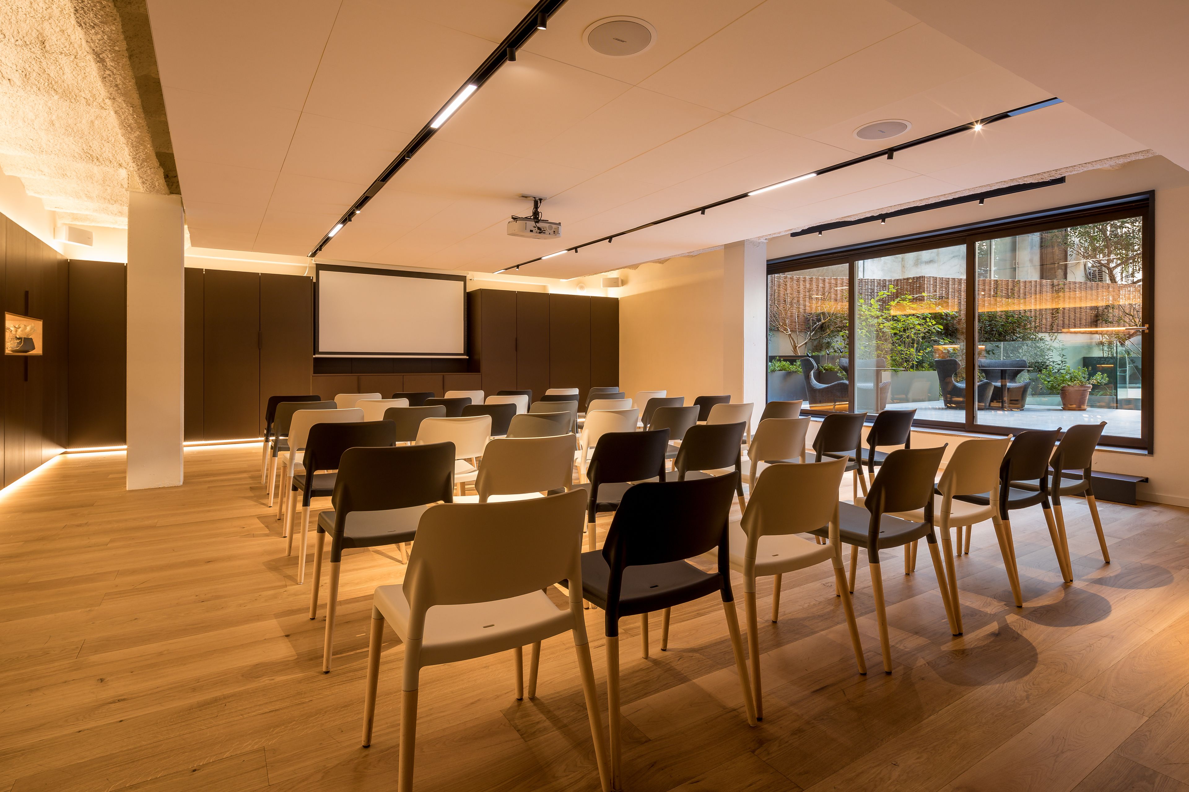 Un salón de conferencias con filas de sillas, una pantalla de proyector y amplias ventanas que dan a vistas del exterior.