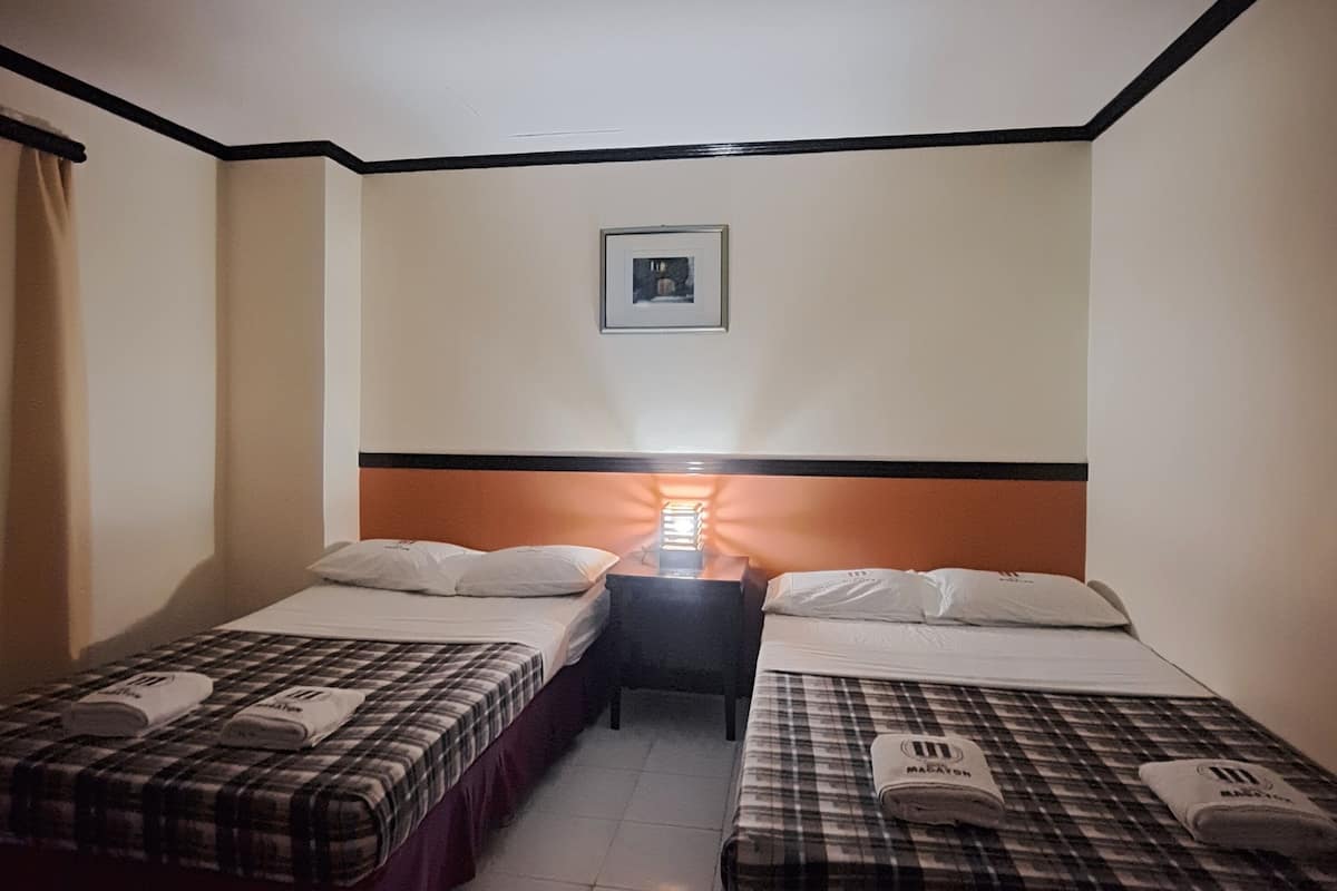 A hotel room with two beds, each with a checkered bedspread and pillows. There is a nightstand between the beds with a lamp and a framed picture on the wall above.