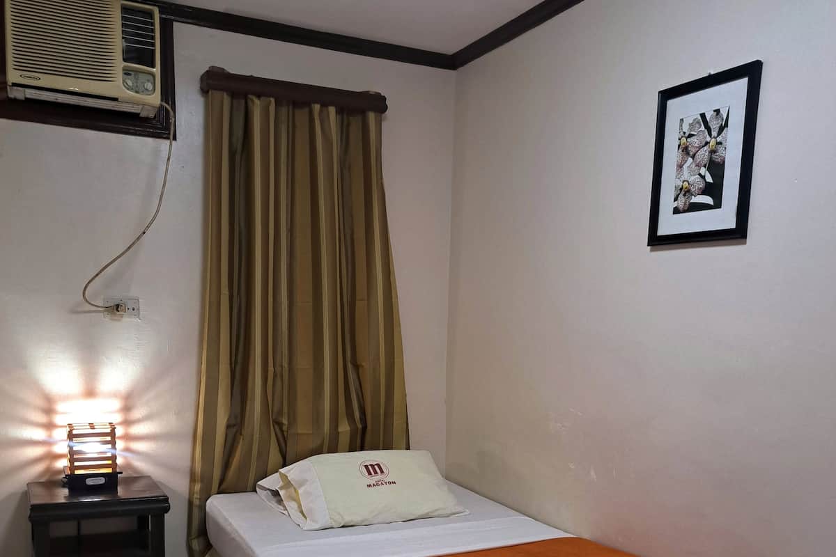 A single bed with a white pillow and a blanket with a logo, a small bedside table with a lamp, a window with curtains, and a framed picture on the wall.