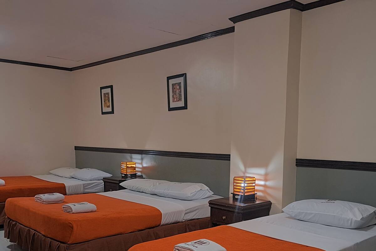A hotel room with three beds, each with an orange bedspread and white pillows. There are two wall-mounted picture frames and a nightstand with a lamp.
