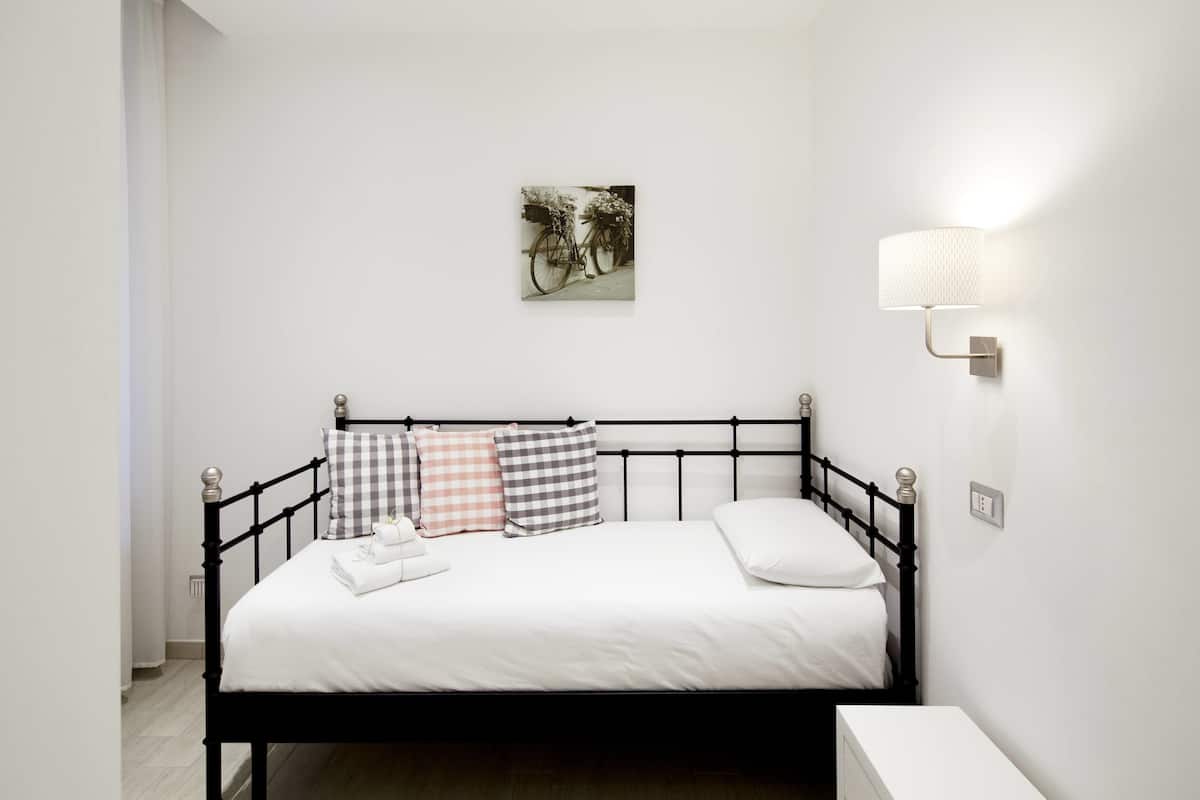 A neatly made bed with a white mattress and black metal frame, accompanied by a variety of checkered pillows. A wall-mounted light fixture is visible to the right of the bed.