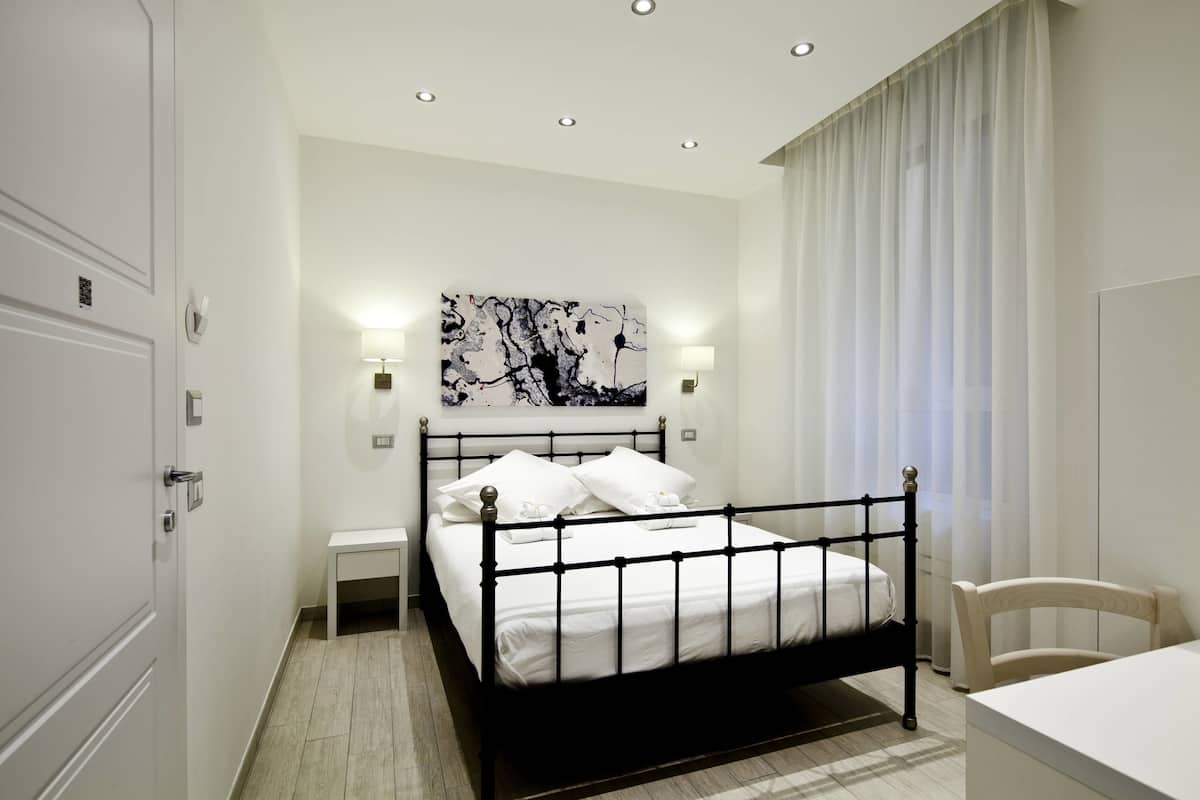 A modern bedroom with a black metal bed frame, white bedding, a bedside table with a lamp, and a large abstract painting on the wall.