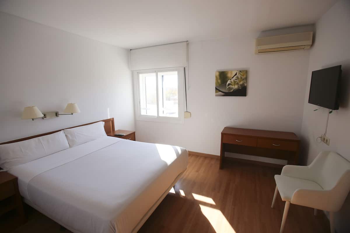 Een slaapkamer met een groot bed, nachtlampjes, een televisie, een raam en een houten dressoir.
