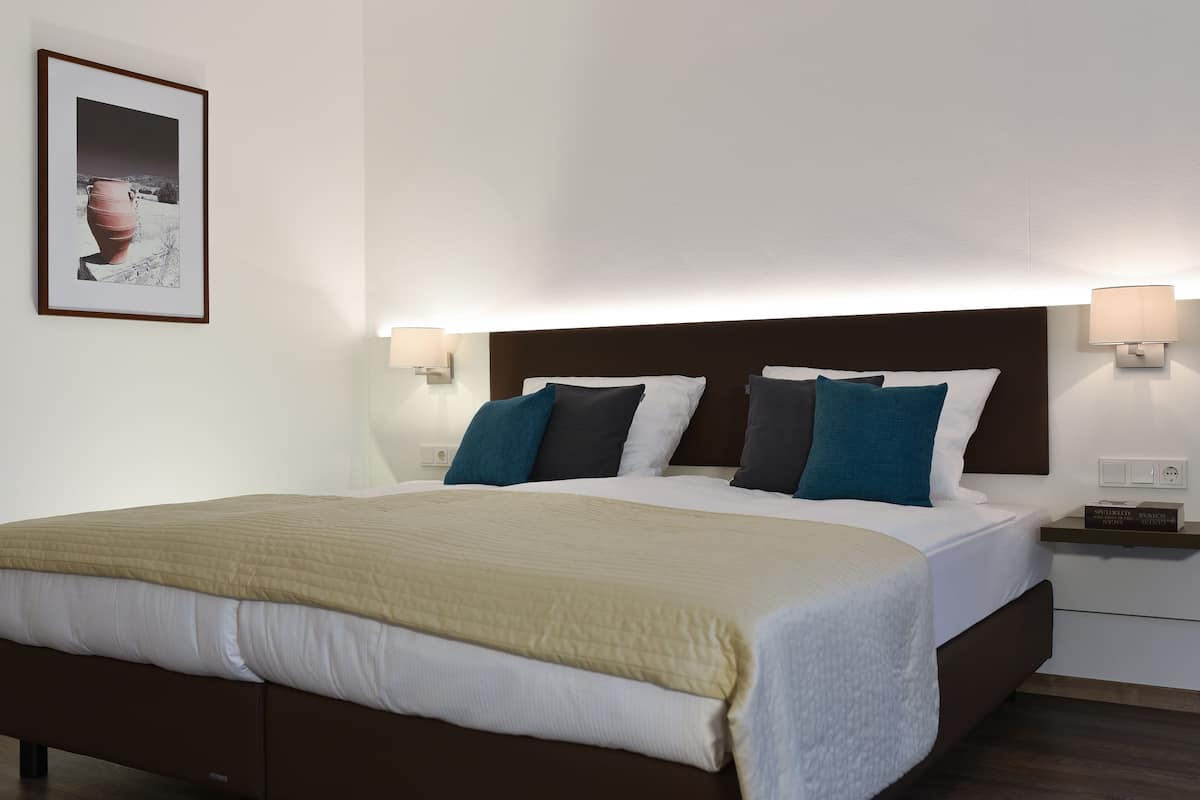 A neatly made bed with a brown headboard, white bedding, and teal pillows. There are two bedside tables with lamps and a framed picture on the wall.