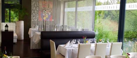 A modern dining area with a black booth and multiple tables set for dining, large windows with a view of greenery, and a decorative curtain.