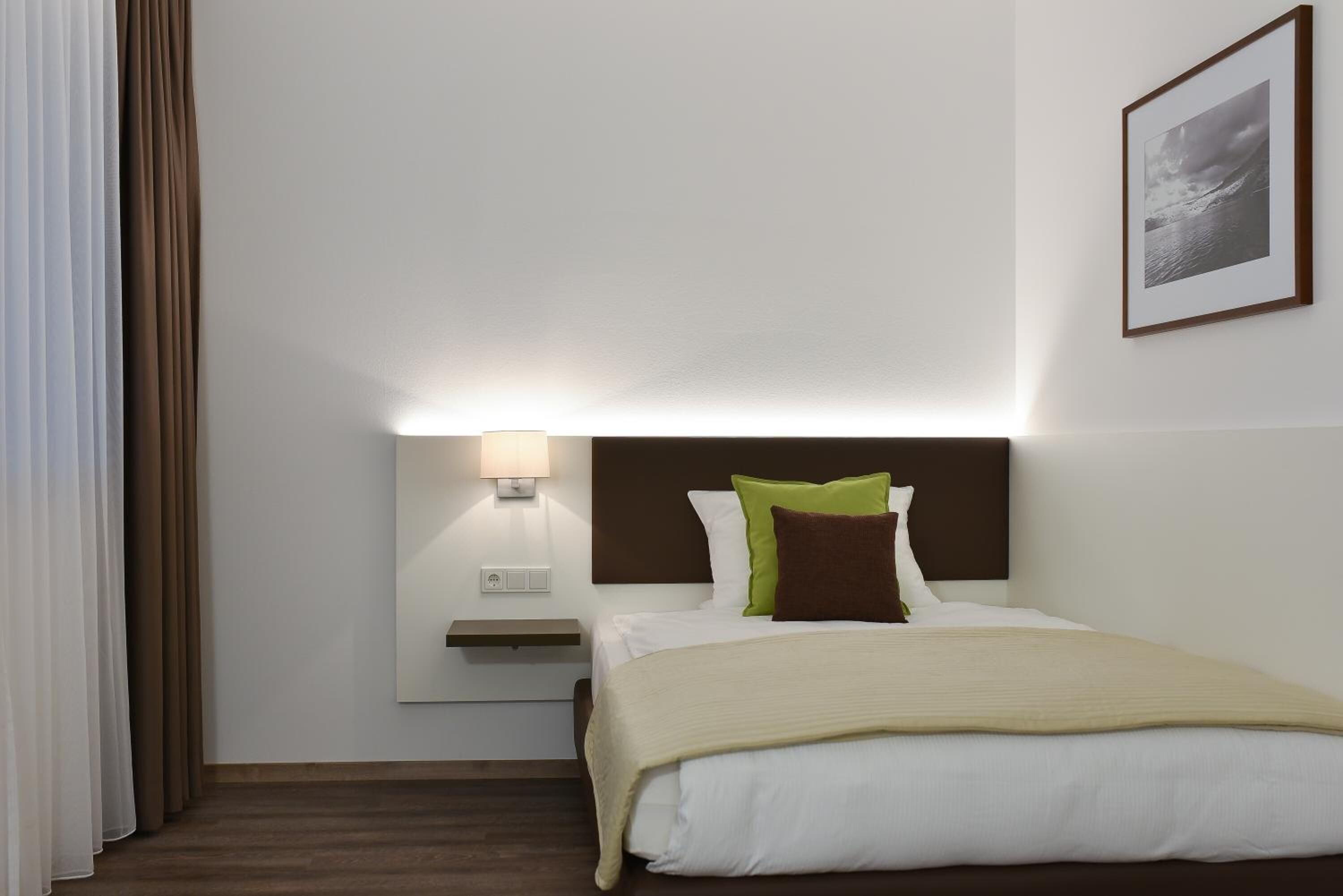 A hotel room with a bed, a nightstand, a wall-mounted lamp, and a framed picture on the wall.