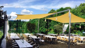 An outdoor dining area with tables and chairs, shaded by a large canopy, set against a backdrop of trees and a body of water. - Guesthouse Lichtenau (Nussloch)
