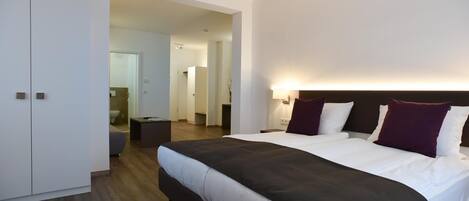 A modern hotel room with a large bed, a bedside table, a wardrobe, and a bathroom visible through an open door.