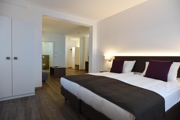 A modern hotel room with a large bed, a bedside table, a wardrobe, and a bathroom visible through an open door.