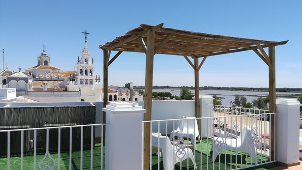Vista de una iglesia blanca con un campanario, una pérgola de madera y un área cercada con sillas blancas.