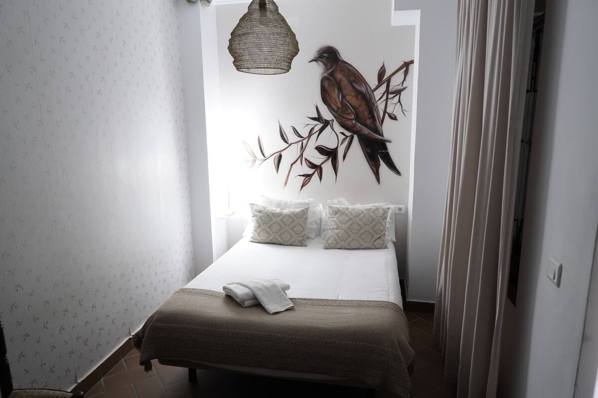 Ein ordentlich bezogenes Bett mit einer braunen Decke und zwei Kissen, ein Wandbild mit Vogelmotiv und eine hängende Leuchte.