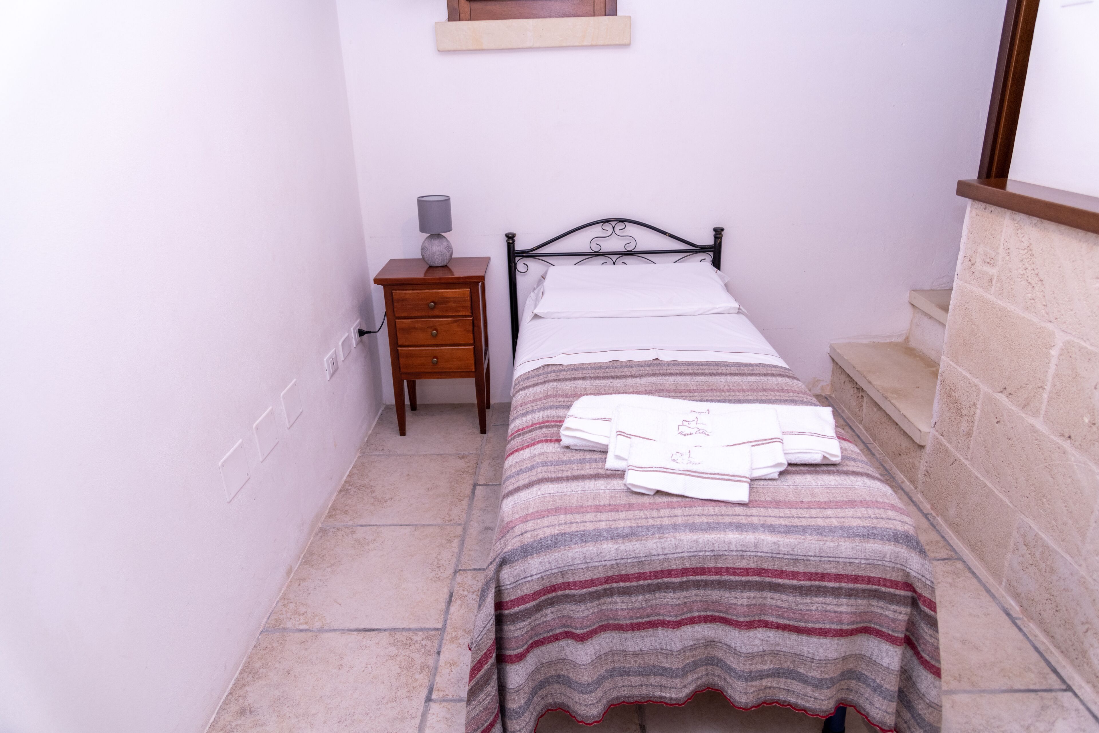 A small, narrow room with a single bed, a nightstand, and a lamp.